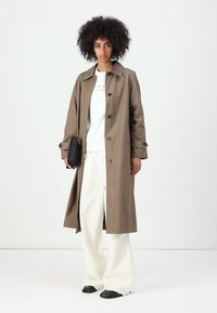 Trench coat beige con vestibilità rilassata, caratterizzato da grandi bottoni, tasche laterali e una texture liscia, indossato sopra una maglietta bianca e pantaloni bianchi larghi.