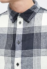 Koszula flanelowa w szaro-białą kratę, charakteryzująca się miękką teksturą, zapinana na guziki, z czarnym kołnierzykiem.