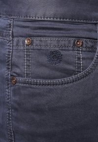 Ciemnoniebieskie jeansy z denimu z przednią kieszenią ozdobioną miedzianymi guzikami i wyszywanym logo, prezentujące subtelne detale szwów.