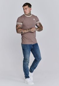 Hnedé tričko s krátkymi rukávmi a bielymi pruhmi na rukávoch a logom, v kombinácii s modrými rifľovými nohavicami a bielymi teniskami, stojaci postoj.