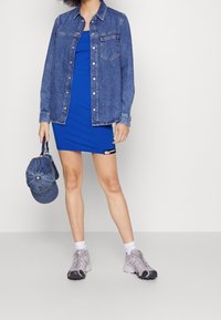 Farkkupaita sinisen istuvan mekon päällä, yhdistetty siniseen lippikseen ja harmaihin urheilukengisiin, korostaen rentoa kerrospukeutumista.