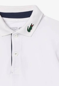 Valkoinen polo-paita tummansinisellä kauluksella, kahdella napilla ja vihreällä krokotiililogo, jossa on punaisia korostuksia vasemmalla puolella. Pehmeää, teksturoitua kangasta.