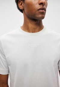 Svetlo šedé tričko s krátkym rukávom vyrobené z mäkkého materiálu, s okrúhlym výstrihom a bielym potlačeným logom značky na prednej strane.