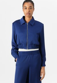 Veste cropped bleu marine avec une fermeture éclair, grand col, poches avant et taille élastique. Texture lisse avec une coupe décontractée.