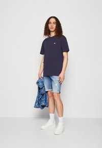 T-shirt di cotone blu navy con un piccolo logo, abbinato a pantaloni corti in denim chiaro sbiancati. Sneakers bianche, giacca di jeans appoggiata al braccio.