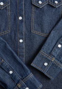 Temna modra denim srajca z bisernimi zapirali, strukturirano yoko obliko in kontrastnim rumenim šivanjem ter dvema žepoma na prsih.