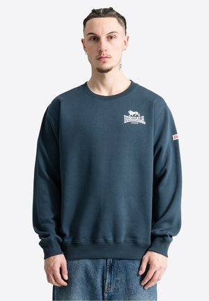 Junger Mann mit geflochtenem Haar, der einen dunkelblauen Lonsdale London Sweatshirt und blaue Jeans vor hellem, einfarbigem Hintergrund trägt.