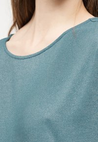 Κοντινό πλάνο ενός ατόμου που φοράει μπλουζάκι σε τιρκουάζ χρώμα με υφή υφάσματος και στρογγυλή λαιμόκοψη, εμφανίζοντας τμήμα του λαιμού και της κλείδας.