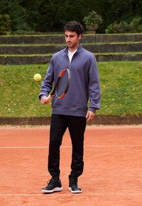 Mladý muž odráža tenisový balón na rakete na antukovom zápasisku s trávnatými schodmi a zeleňou v pozadí.