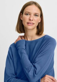 Pull bleu en tricot avec un large col et des manches longues. Le tissu semble doux avec un léger éclat, présentant des poignets côtelés et une coupe décontractée.