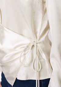 Πουκάμισο από κρεμ σατέν με δέσιμο στο πλάι, που διαθέτει ομαλή υφή και δομημένη σιλουέτα, σε συνδυασμό με σκούρο τζιν.
