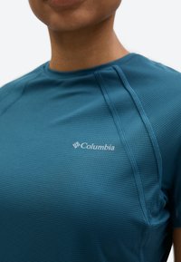 Κοντινό πλάνο ενός ατόμου που φορά μπλε αθλητικό πουκάμισο Columbia με υφή, εμφανείς ραφές και λογότυπο στο στήθος.