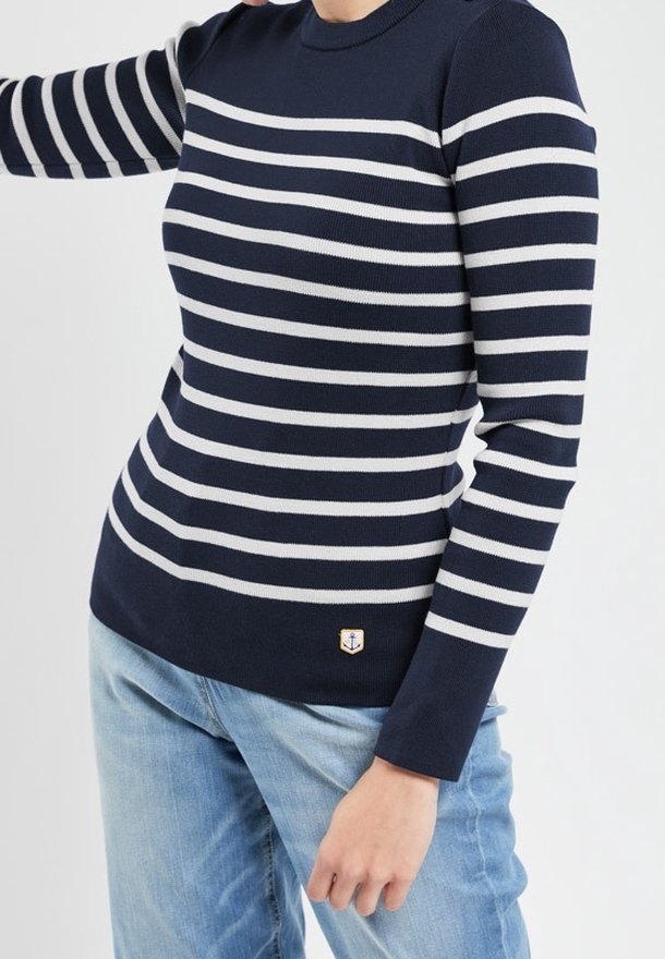 Personne portant un pull rayé horizontal bleu marine et blanc à col rond avec un jean bleu clair sur un fond uni.