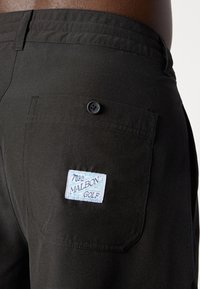 Sorte golfshorts lavet af glat stof, med en baglomme med knap og et lyseblåt mærke med teksten "M.G.C. MALBON GOLF."