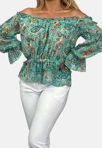 Turkos off-shoulder-blus med paisleymönster, volangkrage och breda ärmar, tillsammans med vita, tajta byxor.