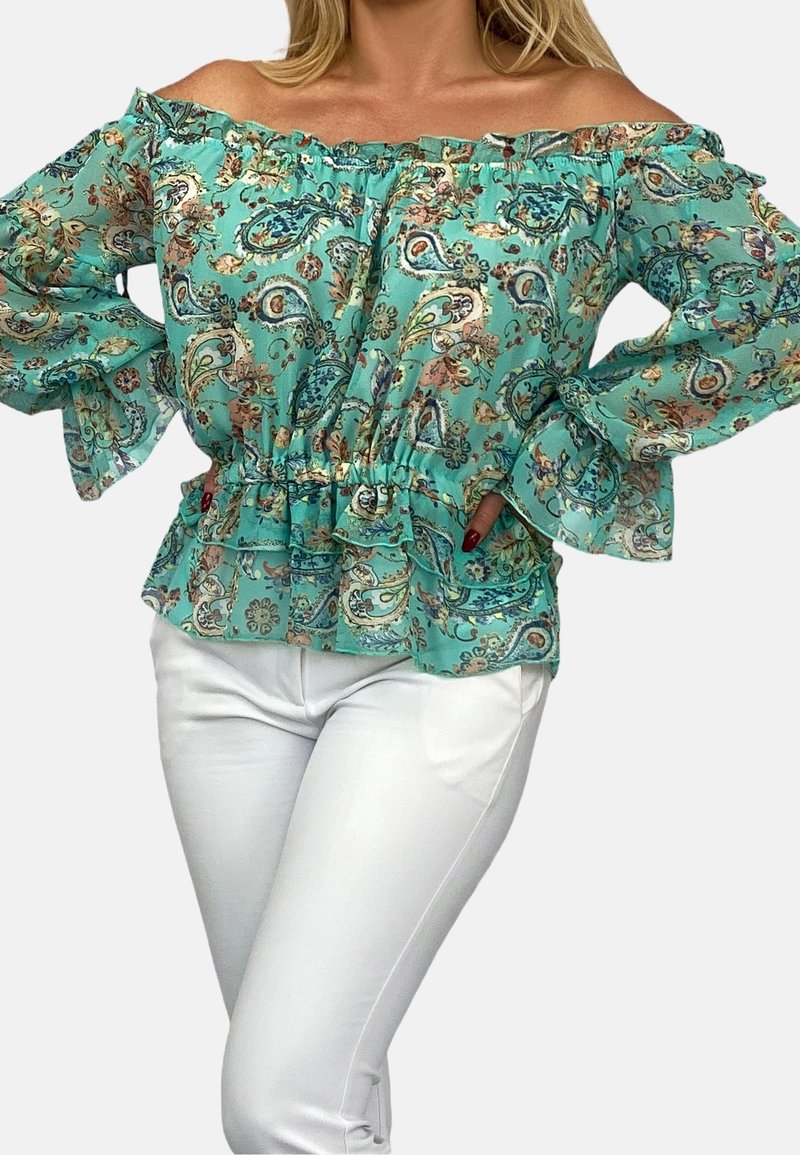 Turkos off-shoulder-blus med paisleymönster, volangkrage och breda ärmar, tillsammans med vita, tajta byxor.