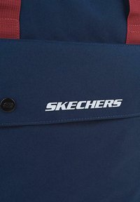 Mochila de tela azul marino con correas rojas y un botón circular negro. Logo blanco prominente de "SKECHERS" en el panel frontal.