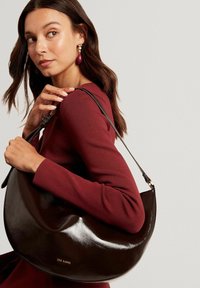 Frau in einem burgunderroten Langarmshirt, die eine glänzende dunkelbraune Schultertasche von Ted Baker hält und über ihre linke Schulter blickt.