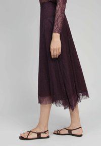 Jupe midi en dentelle de couleur violet profond avec un design évasé et un ourlet scallopé. Associée à des sandales plates marron avec des bretelles fines et une boucle décorative.