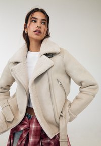 Beige Schaffelljacke mit breitem Kragen, seitlichem Reißverschluss- oder Stecktasche und passenden Bündchen. Über einem weißen Hemd getragen, mit einem Karo-Tuch um die Taille gebunden.