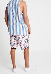 Débardeur pour homme avec des rayures verticales bleues et blanches, associé à un short à motif floral en blanc, rose et bleu, avec des accents noirs.
