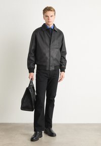 Melnā ādas bomber jaka ar ribotām aprocēm, ar izceltu logo. Saskaņota ar melnajiem džinsiem un gludām melnām zābakiem, tur rokās melnu somu.