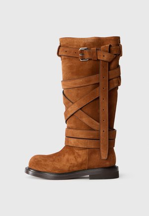 Botte en cuir suédé marron, montante jusqu'au genou, avec des sangles croisées, deux boucles en argent et une semelle plate. Présente une pointe arrondie et une surface texturée.