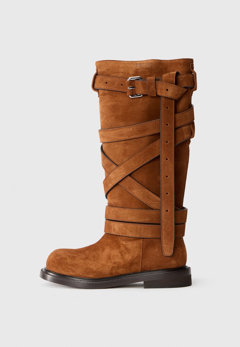 Brown suede knee-high boot with crisscross straps, two silver buckles, and flat sole. Features a rounded toe and textured surface.
