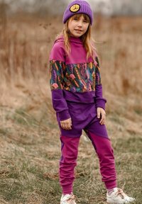 Fioletowy dres z kolorową, liściastą górną częścią; posiada kaptur, elastyczne mankiety oraz pasujące spodnie dresowe. Noszony z fioletową czapką beanie.