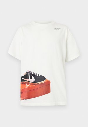 Biała koszulka z krótkim rękawem z nadrukiem czarnego korka piłkarskiego Nike na zużytym czerwonym pudełku na buty w pobliżu dolnej krawędzi oraz małym logo przy dekolcie.