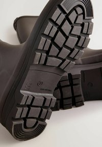 Bottes en caoutchouc noires avec des semelles texturées et durables, présentant un motif de semelle profond. Matériau supérieur lisse, bout renforcé et logo latéral.