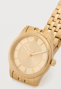 Αναλογικό ρολόι χειρός σε χρυσό τόνο με λογότυπο BOSS, ρωμαϊκό αριθμό XII και χρυσό μπρασελέ με κρίκους, δείχνει την ώρα 10:10:36.