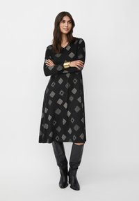 Robe noire à manches longues, ornée de motifs géométriques en argent. Texture lisse, décolleté en V, accompagnée de bottes noires jusqu'aux genoux.