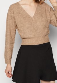 Haut en tricot beige à manches longues, en croisé, avec une finition texturée et un décolleté en V profond, associé à une jupe noire fluide.