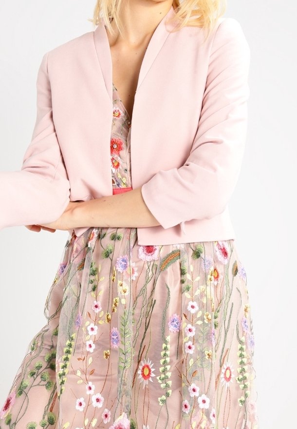 Lichtroze cropped jack met een sjaalkraag en lange mouwen. Gedragen over een jurk met bloemenborduursels in pastelkleuren en fijne details.