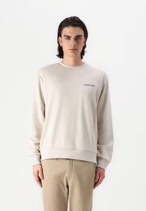 Beige bomullssweatshirt med rund hals, ribbestrikkede mansjetter og nederkant. Har teksten "Club1990" i svart på brystet. Brukt med beige bukser.