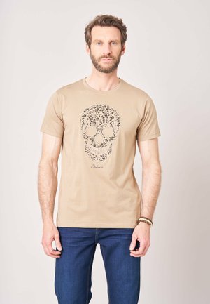 Homme barbu portant un t-shirt beige avec un motif de crâne floral et un jean bleu, debout devant un fond uni.