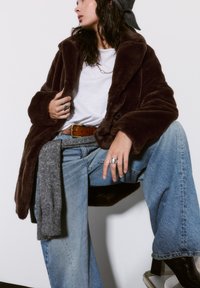 Rjav plašč iz umetnega krzna s širokimi reverji, gumbom za zapiranje in globokimi sprednjimi žepi. V kombinaciji s sproščenimi modrimi kavbojkami in sivo ruto.