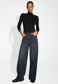 Femme se tenant avec les mains dans les poches, portant un col roulé noir à manches longues, un jean large foncé et des chaussures noires sur fond blanc.