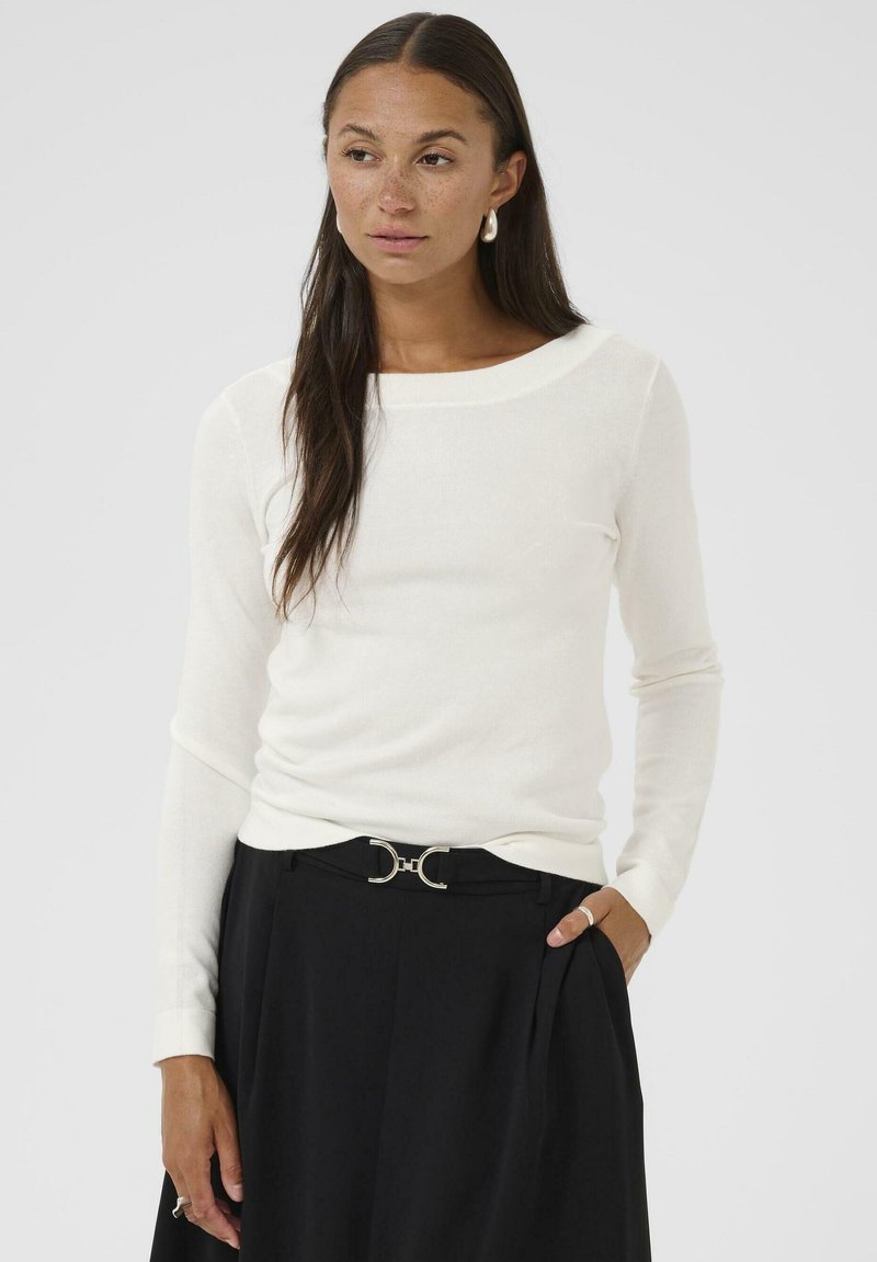 Pull long à manches blanches avec un col rond, fabriqué dans un matériau doux. Associé à un pantalon noir taille haute avec une ceinture à accent métallique.