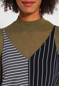 Randig svart och vit klänning med V-ringning över en olivgrön polotröja, med kontrasterande mönster och texturer i tyget.