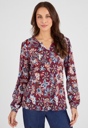 Lange mouw blouse in een wijnrode stof met bloemenpatronen in blauw en wit. Heeft een V-hals en plooidetails bij de halslijn.