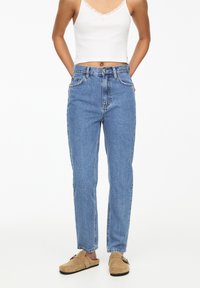 Mujer que lleva unos vaqueros de tiro alto azules, una camiseta corta blanca con acabados de encaje y unos zapatos de ante beige tipo slip-on con detalle de hebilla.