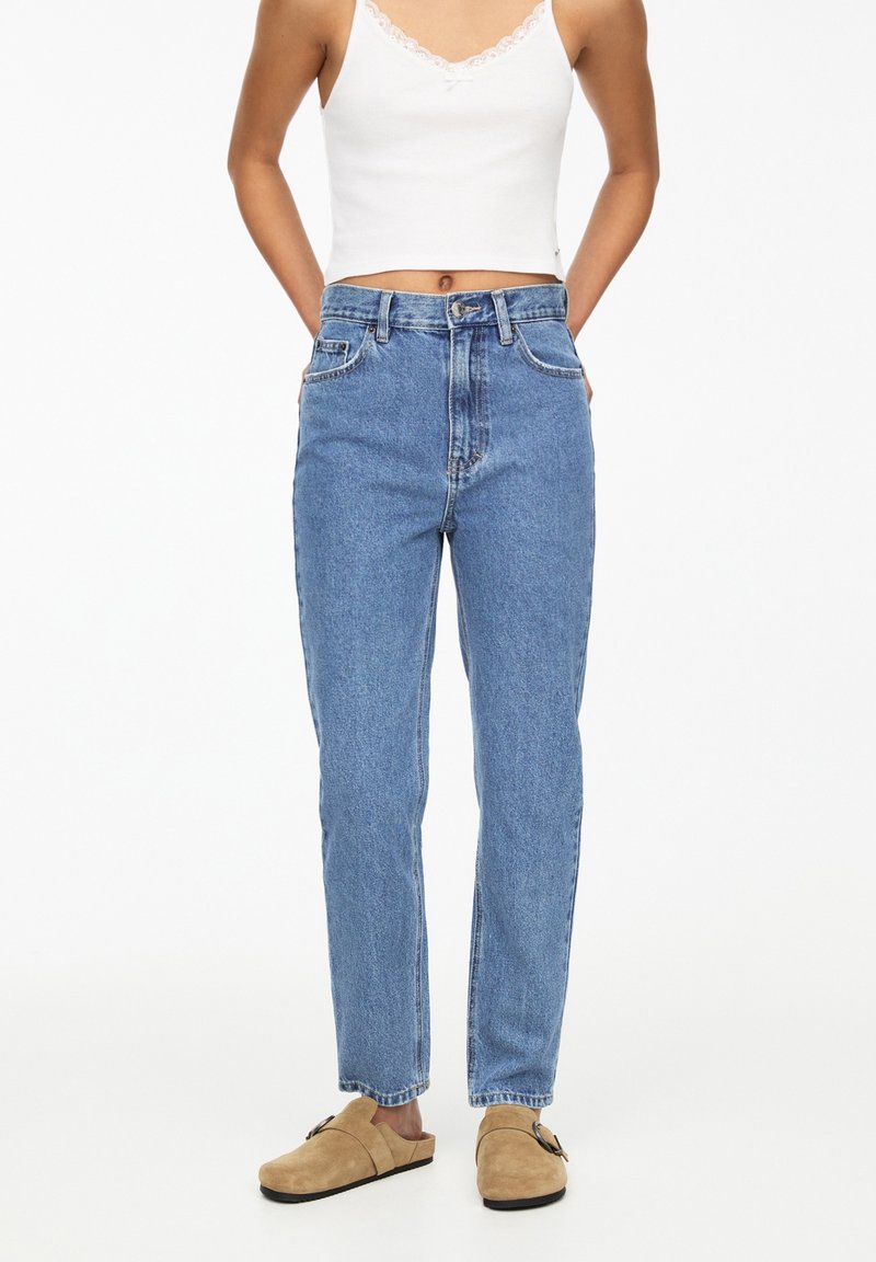 Mujer que lleva unos vaqueros de tiro alto azules, una camiseta corta blanca con acabados de encaje y unos zapatos de ante beige tipo slip-on con detalle de hebilla.