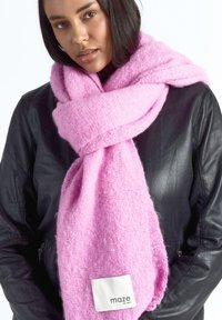 Vrouw draagt een dikke, zachte roze sjaal met het label "maze" om haar nek, over een zwarte leren jas, tegen een witte achtergrond.