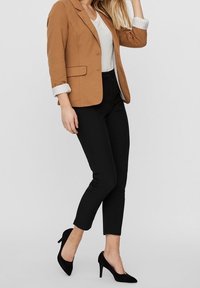 Hnědý blazer s těsným střihem, bílá košile pod ním, černé slim-fit kalhoty a černé stiletto podpatky. Neformální, ale přesto profesionální vzhled.