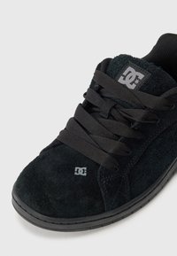 Zwarte suède skate schoen met een gevoerde tong, platte zwarte veters en een grijs logo op de zijkant. Bevat een rubberen zool met een gestructureerd oppervlak.