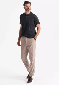 Černé polo tričko se zipovým límcem, doplněné o světle béžové zúžené kalhoty a hnědé šněrovací boty. Model má plnovous a brýle.