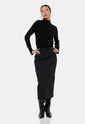 Mujer de pie con las manos en las caderas, vestida con un jersey de cuello alto negro, falda larga negra y botas negras hasta la rodilla sobre fondo blanco.