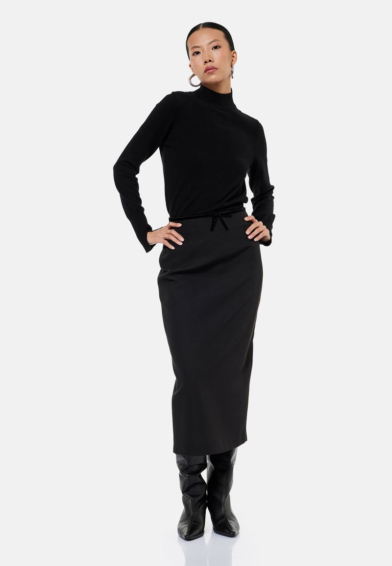 Mujer de pie con las manos en las caderas, vestida con un jersey de cuello alto negro, falda larga negra y botas negras hasta la rodilla sobre fondo blanco.